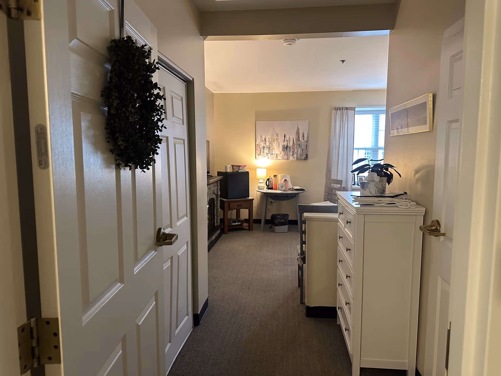 View down a hallway into a cozy living area with a small table, chair, lamp, and a dresser. There is a wreath hanging on the door to the left and a window with curtains letting in natural light. The walls are painted a light beige color and there is artwork on the walls.