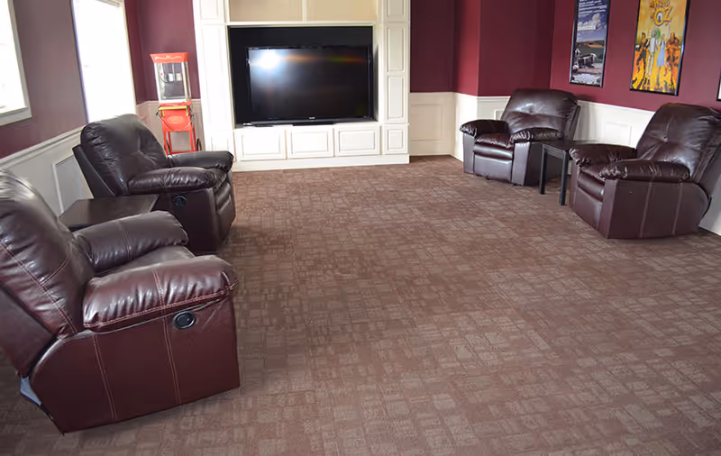 A cozy living room with four dark brown leather recliners arranged around a large flat-screen TV mounted on a white built-in cabinet. The room has maroon walls with white wainscoting and carpeted floor. There is a red popcorn machine in the corner and framed posters on the wall.