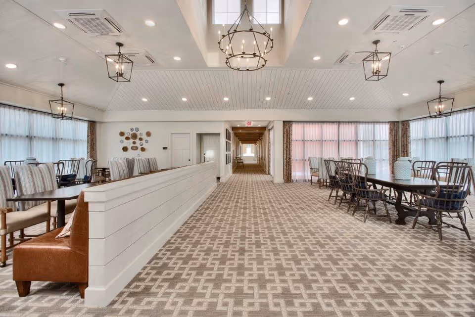 A spacious, well-lit dining area with multiple tables and chairs arranged neatly on a patterned carpet. The room features large windows with sheer curtains allowing natural light to fill the space. Modern chandeliers hang from the ceiling, and a long white half-wall divides the seating areas. The far end of the room shows a hallway with an exit sign above the door.