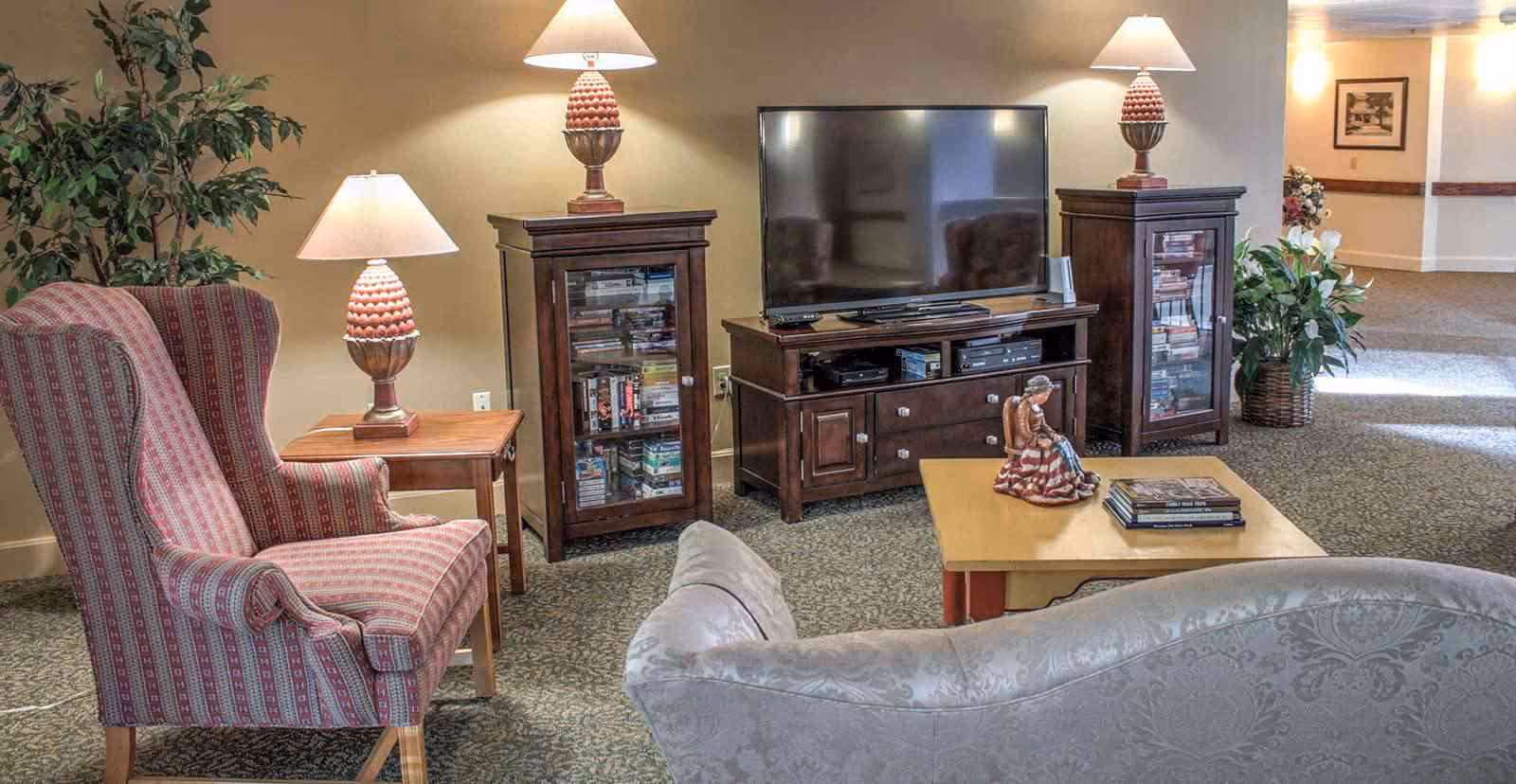 A cozy living room area with a patterned armchair, a sofa, a wooden coffee table with a figurine and books, two wooden cabinets with glass doors holding books and DVDs, a flat-screen TV on a wooden TV stand, and three table lamps providing warm lighting. There are also plants and framed artwork on the walls.