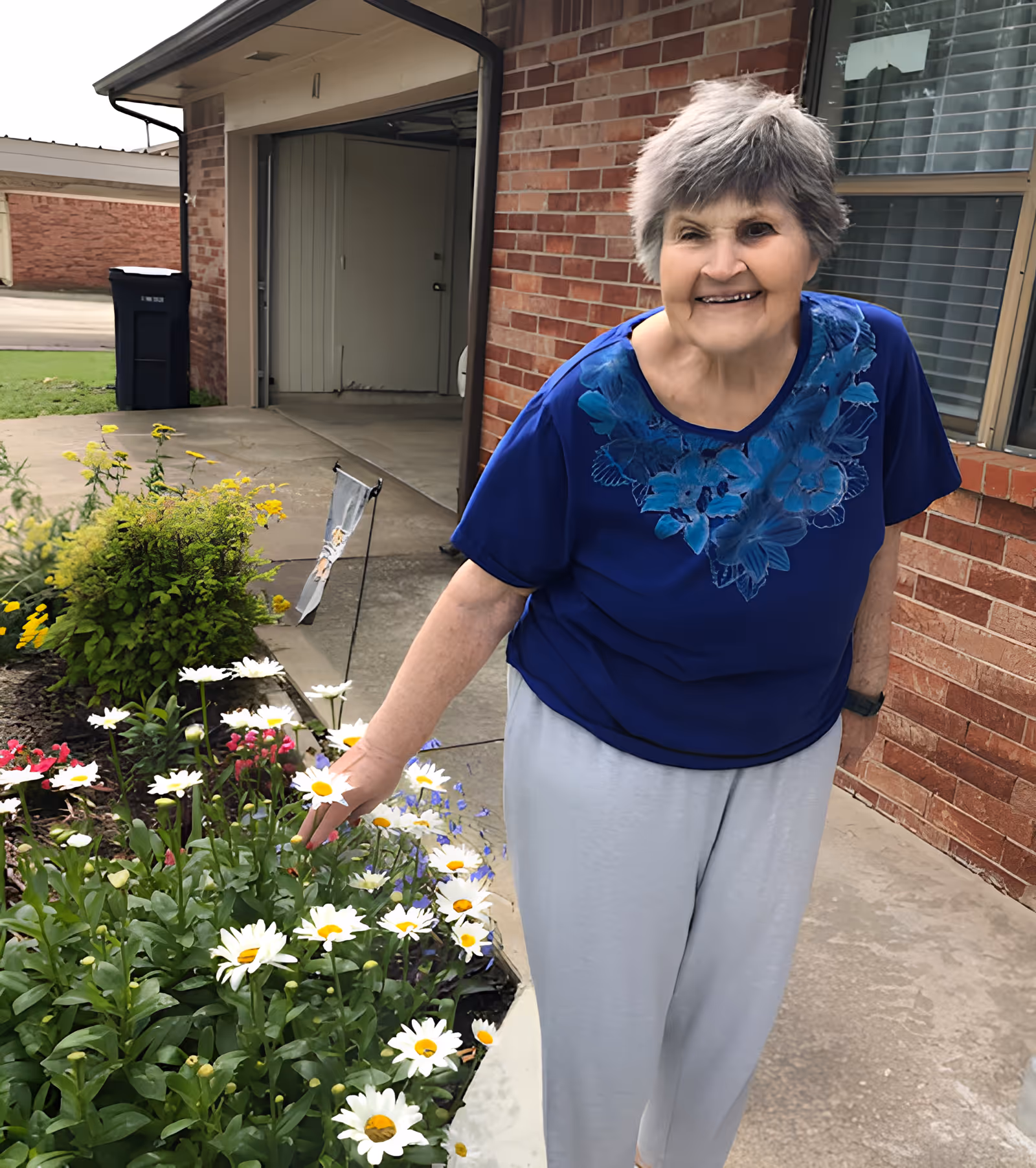 An elderly woman wearing a blue shirt with floral designs and light gray pants is standing outside near a flower bed with white daisies and other colorful flowers. She is smiling and gently touching one of the daisies. Behind her is a brick building with a window and an open garage door.