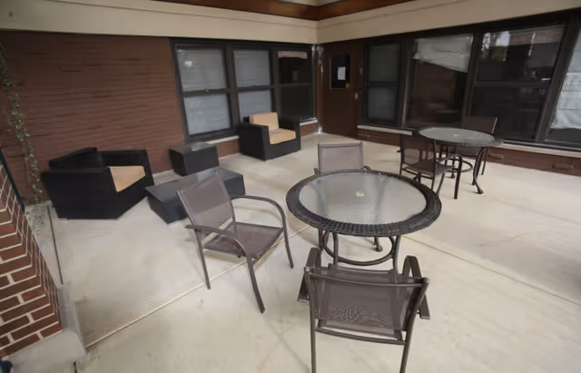 Covered outdoor patio area with two round glass-top tables and several chairs arranged around them. There are also cushioned armchairs and small tables against a brick wall with windows and a door leading inside.