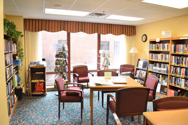 A cozy library room with bookshelves filled with books along the walls, a central table surrounded by six maroon upholstered chairs, a patterned blue carpet, large windows with striped valances letting in natural light, and a floor lamp in the corner.