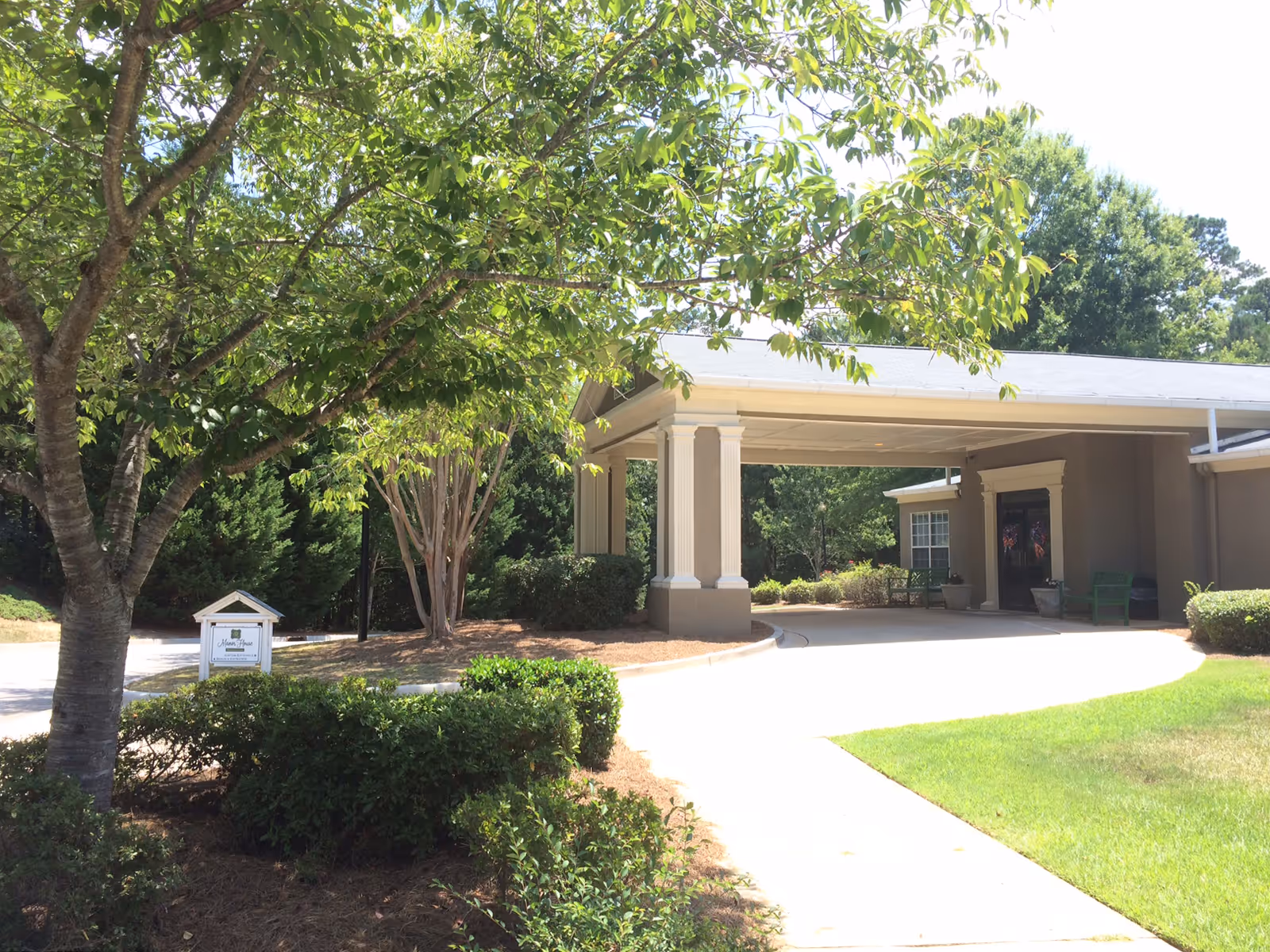 Exterior view of a senior living facility entrance with a covered driveway, surrounded by green trees and bushes under a sunny sky.