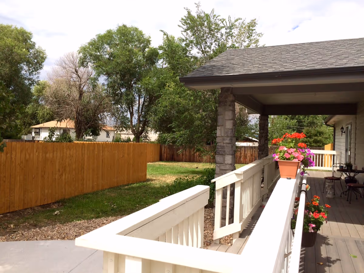 A covered porch area with a white wooden railing and a ramp leading down to a concrete path. There are flower pots with colorful flowers placed along the railing. The porch is supported by stone pillars and overlooks a fenced backyard with green grass and trees.