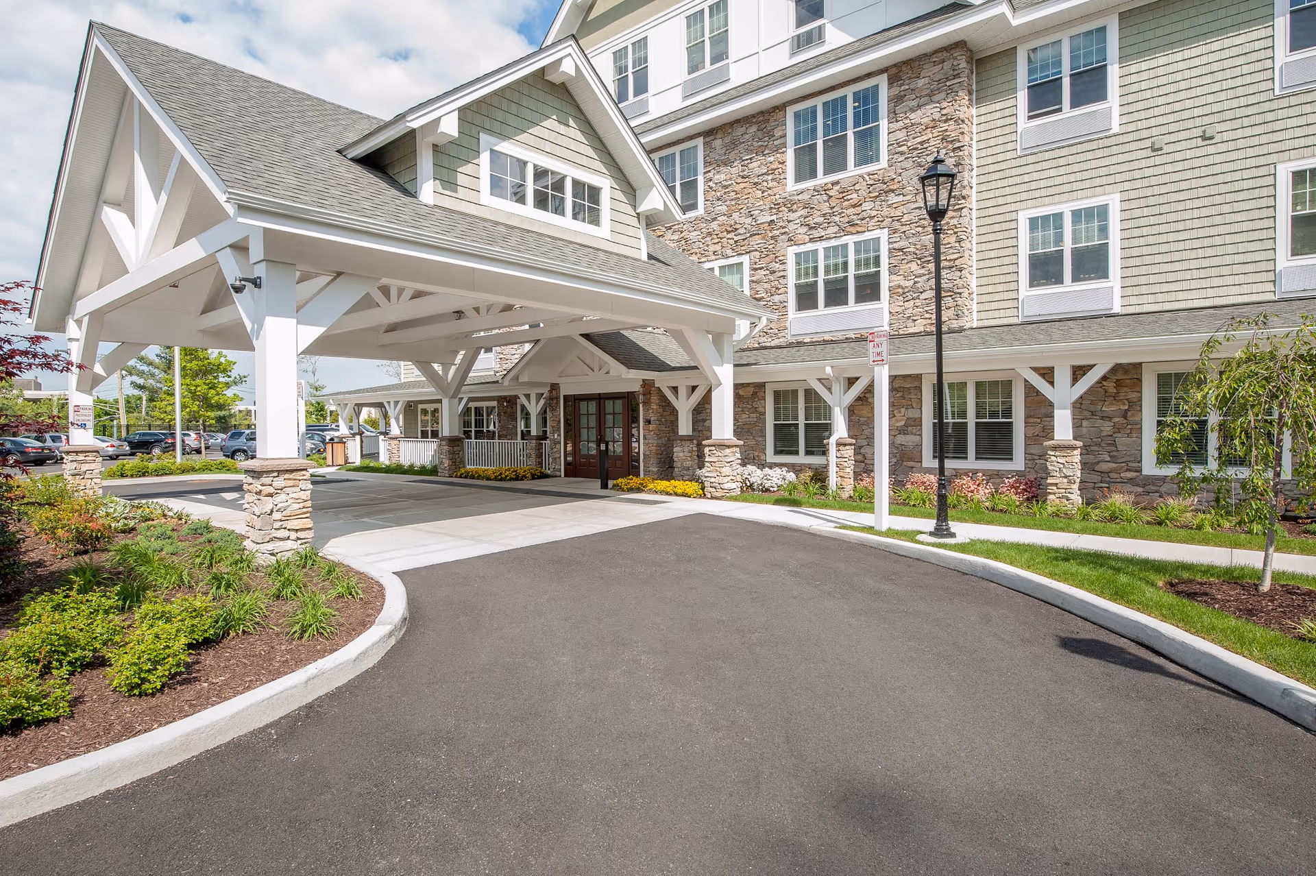 Entrance of a senior living facility with a covered driveway supported by white wooden beams and stone pillars. The building exterior features a combination of stone and green siding with multiple windows. There are landscaped plants and a lamppost near the entrance, and a parking lot is visible in the background under a partly cloudy sky.