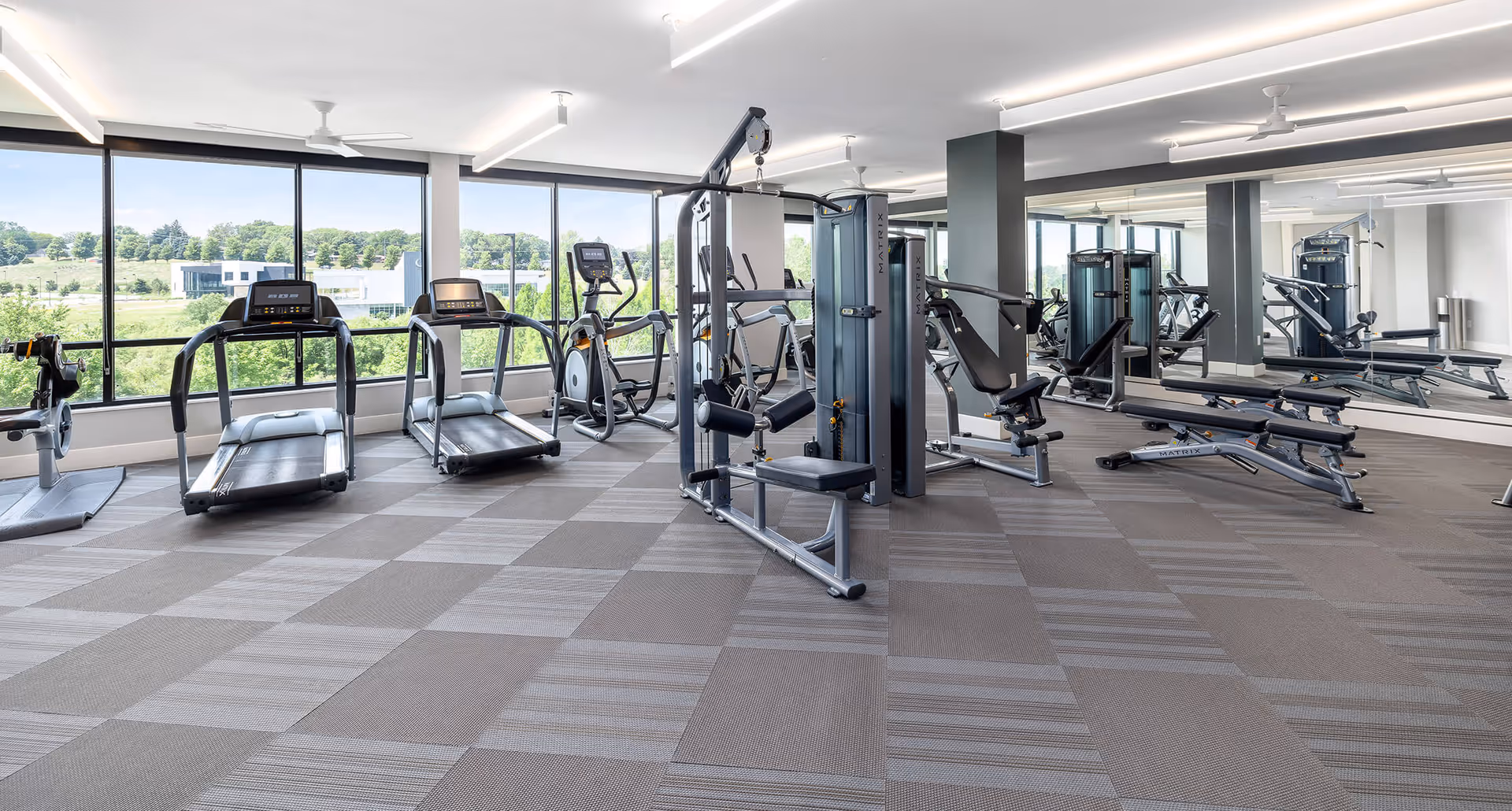 Bright and spacious fitness center with large windows showing a green outdoor view. The room contains various exercise equipment including treadmills, elliptical machines, weight machines, and benches. The floor is covered with patterned carpet tiles, and one wall is covered with mirrors.