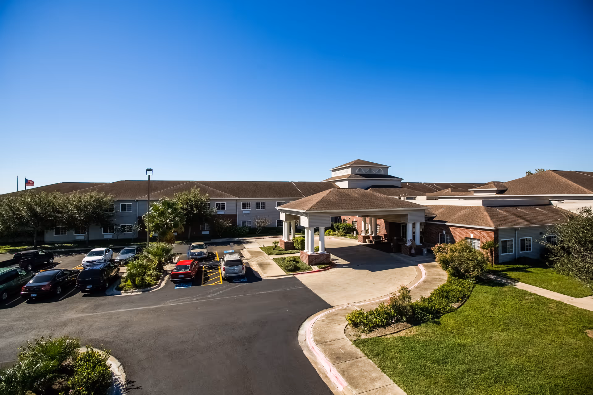 Exterior view of Garden Estates of Corpus Christi senior living facility showing a large building with a covered entrance, parking lot with several cars, landscaped greenery, and a clear blue sky.