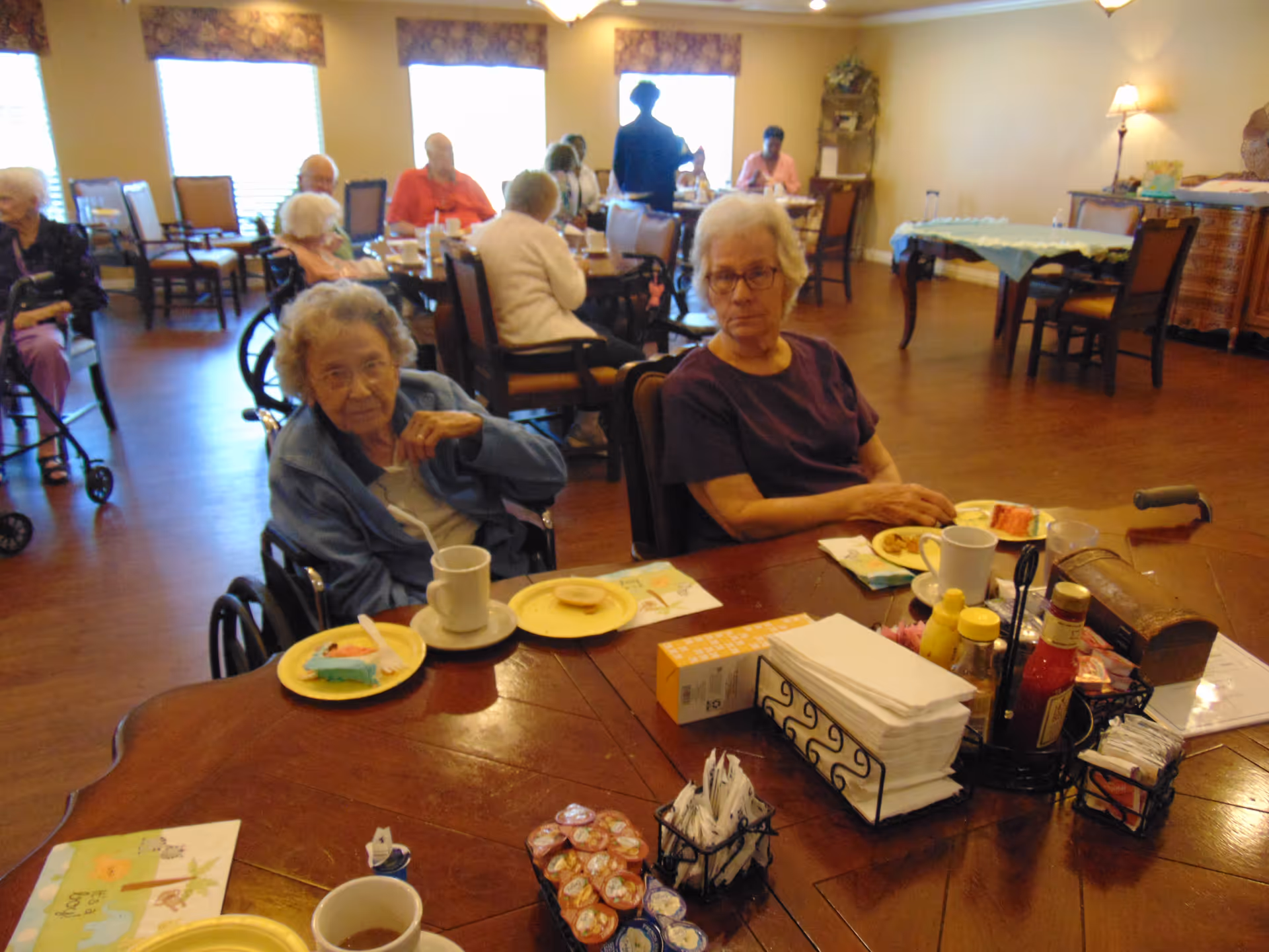 A dining room in a senior living facility with elderly residents seated at tables eating and drinking. Two elderly women are seated at the foreground table with plates of food and cups. Other residents and staff are visible in the background. The room has wooden floors, large windows with floral valances, and warm lighting.