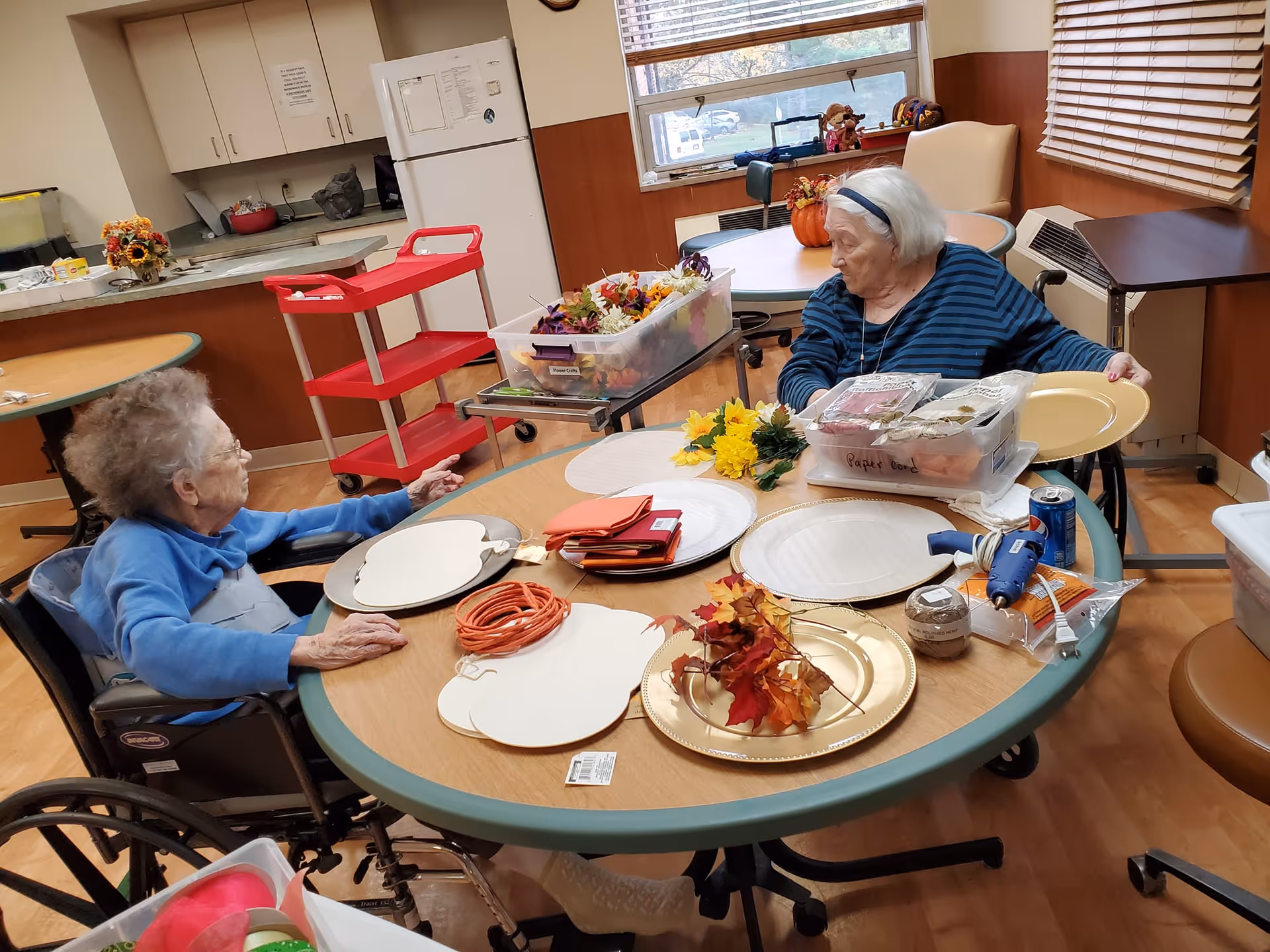 Two elderly women in wheelchairs sitting at a round table in a room with wood flooring and beige walls. The table is covered with craft supplies including artificial flowers, plates, napkins, and a hot glue gun. In the background, there is a kitchen area with a refrigerator, cabinets, and a red utility cart. A window with blinds lets in natural light.