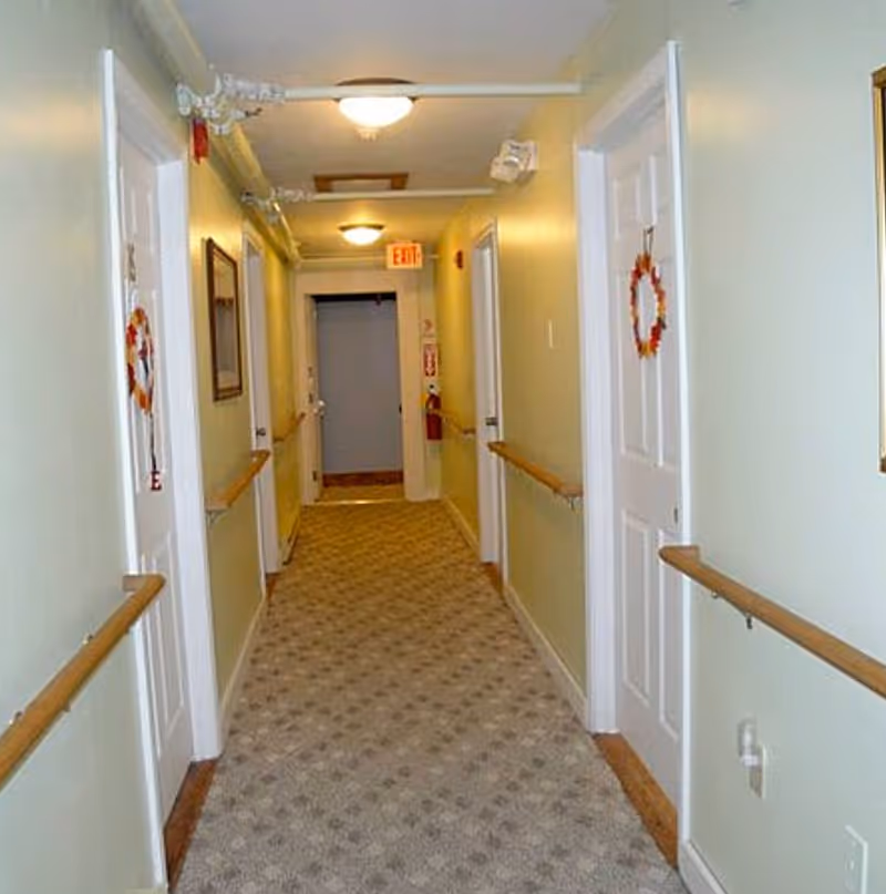 A carpeted hallway in a senior living facility with light green walls, white doors on both sides decorated with small wreaths, wooden handrails along the walls, ceiling lights, and an exit sign at the far end.
