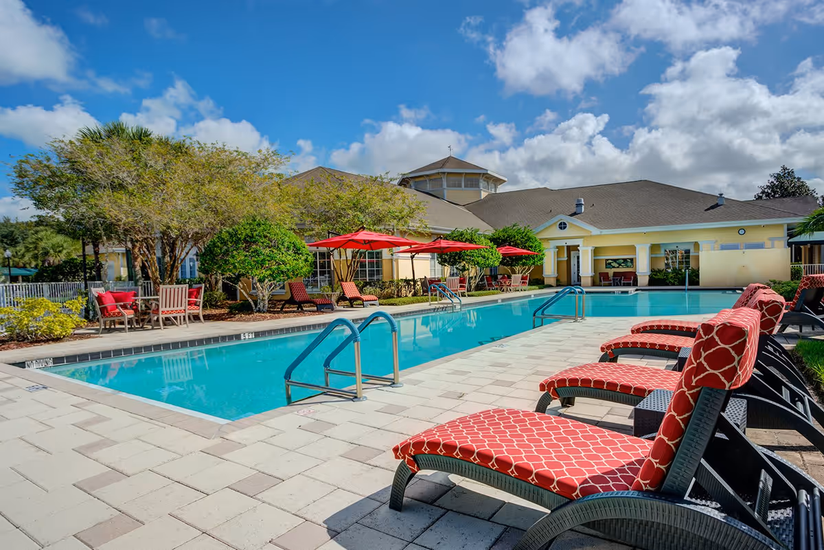 Outdoor swimming pool area with red cushioned lounge chairs and red umbrellas. The pool is surrounded by a tiled deck and greenery, with a yellow building in the background under a partly cloudy blue sky.
