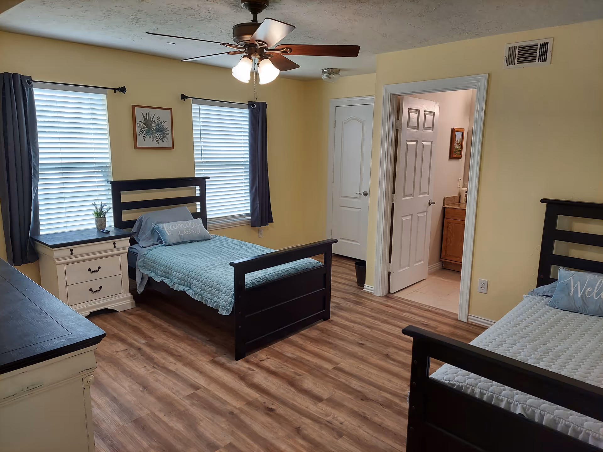 A bedroom with two single beds featuring dark wooden frames and light blue bedding. The room has light yellow walls, wood-patterned flooring, two windows with white blinds and dark curtains, a ceiling fan with lights, a white dresser, and a door leading to a bathroom with a visible sink and cabinet.