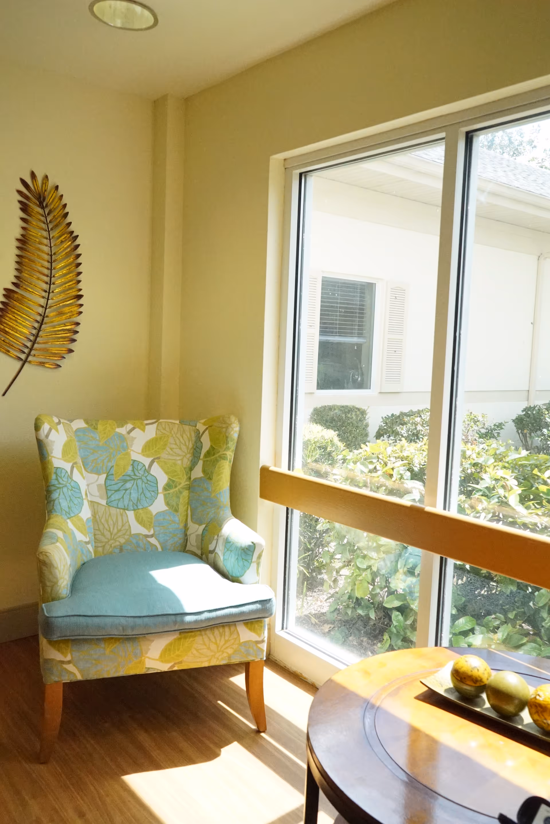 A cozy corner with a floral patterned armchair featuring blue and green leaves, positioned next to a large window letting in natural light. A round wooden table with decorative fruit is partially visible in the foreground. Outside the window, bushes and part of a building with a window and shutters can be seen.