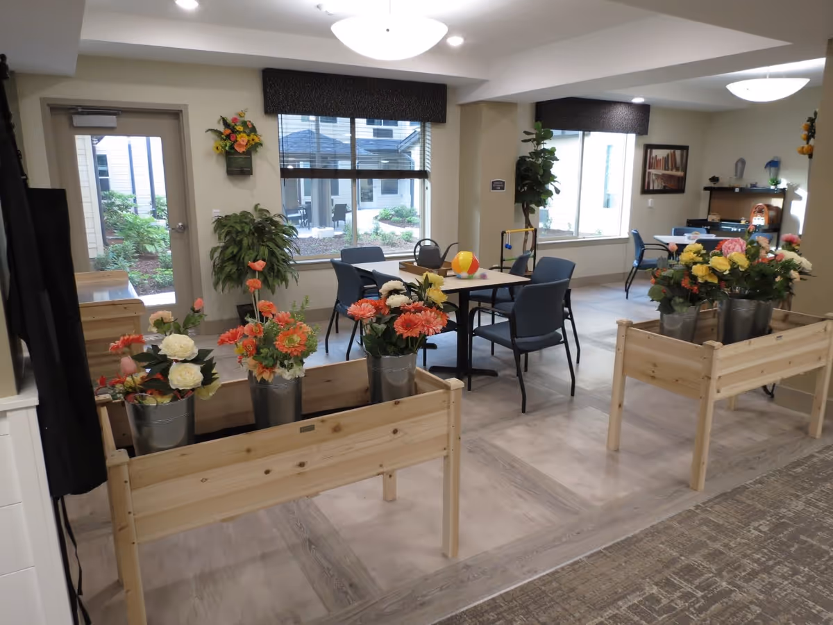 A bright and spacious common area with tables and chairs, decorated with colorful artificial flowers in metal buckets placed on wooden stands. Large windows let in natural light and offer a view of a garden outside. The room has light-colored walls and flooring, with a few plants and framed pictures on the walls.