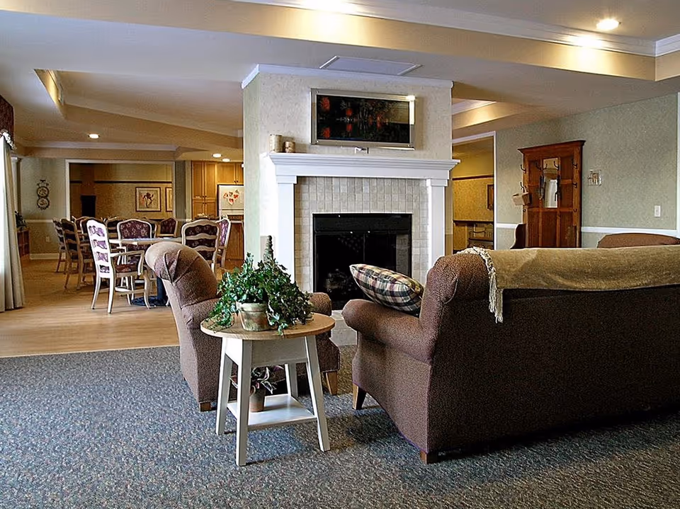 A cozy living room area with two brown upholstered armchairs and a matching sofa arranged around a white fireplace with a decorative painting above it. A small wooden side table with a potted plant sits between the chairs. In the background, there is a dining area with a table and chairs, and a kitchen area further back. The room has soft lighting and a warm, inviting atmosphere.
