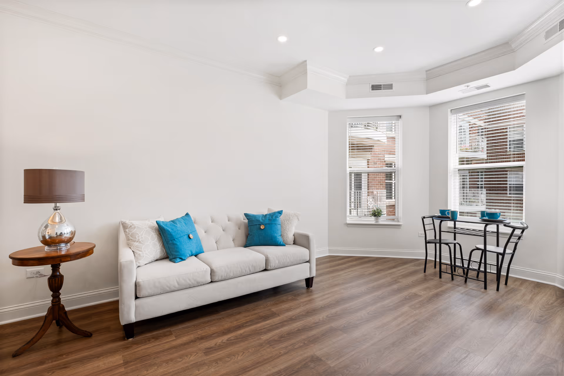 Bright living room with a white sofa accented by turquoise pillows, a wooden side table with a lamp, and a small dining table by bay windows.