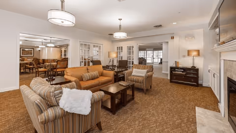 A furnished senior living common room with sofas, armchairs, coffee tables, chandeliers, and a fireplace.