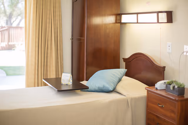 A neatly made single bed with a light beige blanket and a blue pillow in a room. There is a wooden headboard and a small wooden bedside table with a white telephone and a small potted plant. A wooden cabinet is visible behind the bed, and a window with beige curtains lets in natural light. A small tray with a welcome card is placed on the bed.