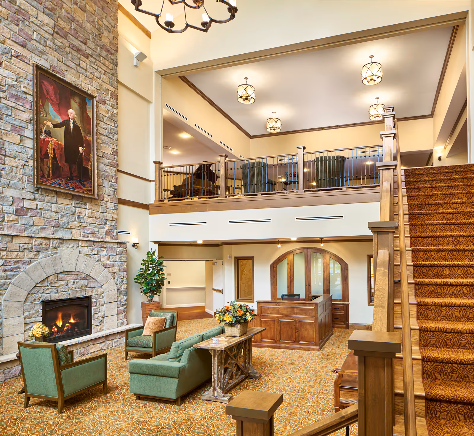 A spacious and warmly lit senior living facility lobby with a stone fireplace, green upholstered chairs and sofa, a wooden reception desk, a staircase with patterned carpet, and an upper balcony area with additional seating and a grand piano. A large portrait hangs above the fireplace.
