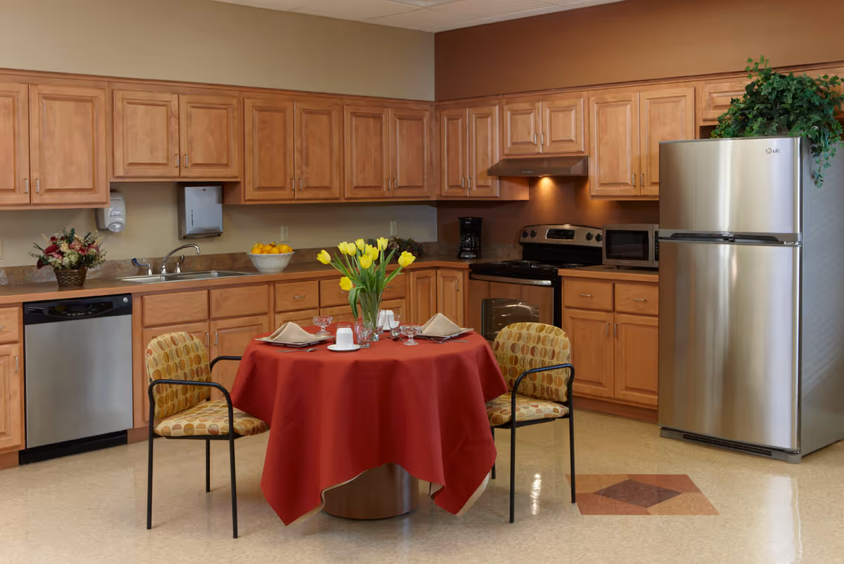 A kitchen area with wooden cabinets, a stainless steel refrigerator, dishwasher, stove, and microwave. In the center is a round table covered with a red tablecloth, set with plates, glasses, napkins, and a vase of yellow tulips. Two chairs with patterned upholstery are placed around the table. There are decorative plants and a bowl of lemons on the countertop.