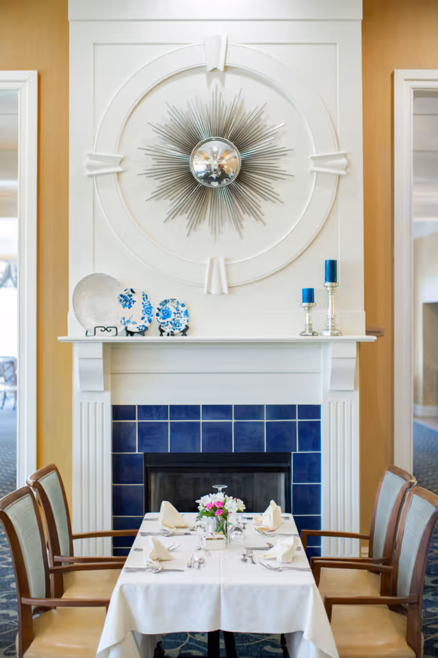 A dining table set for four with white tablecloth, napkins, cutlery, and a floral centerpiece in front of a white fireplace with blue tiles. Above the fireplace is a decorative sunburst mirror and decorative plates and candles on the mantel. The room has beige walls and carpeted floors.