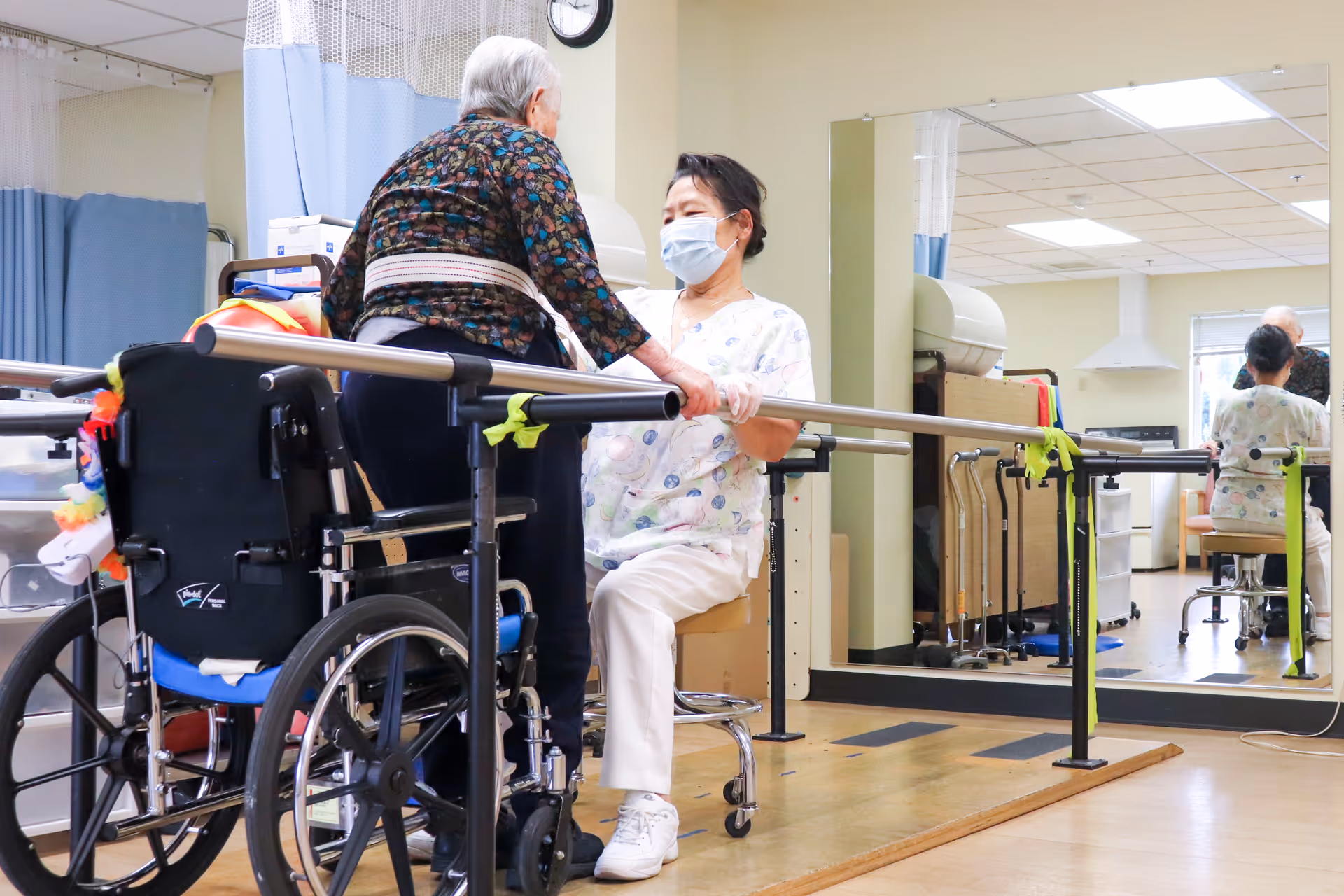 An elderly woman using parallel bars for physical therapy assistance, supported by a healthcare worker wearing a mask in a rehabilitation room with a wheelchair nearby and a large mirror on the wall.