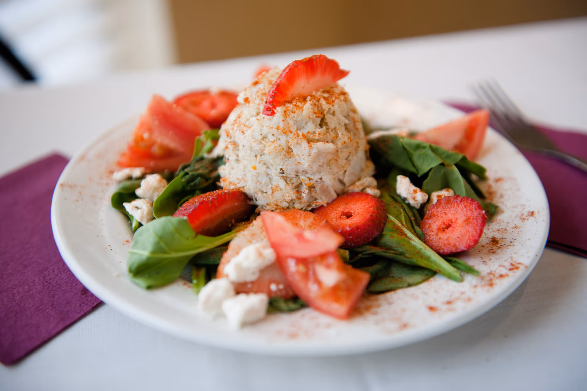 Plate of salad with a molded scoop of crab or chicken salad topped with a sliced strawberry, served on spinach with tomato wedges and crumbled cheese.