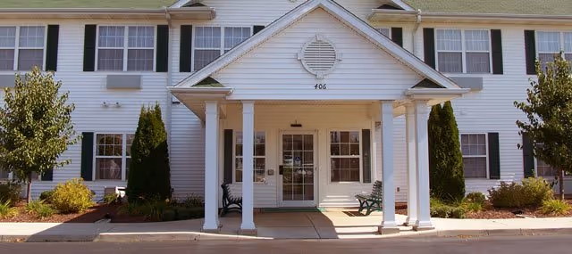 Front exterior view of a two-story white building with multiple windows, a covered entrance supported by four white columns, two benches on either side of the entrance, and landscaping with bushes and small trees.