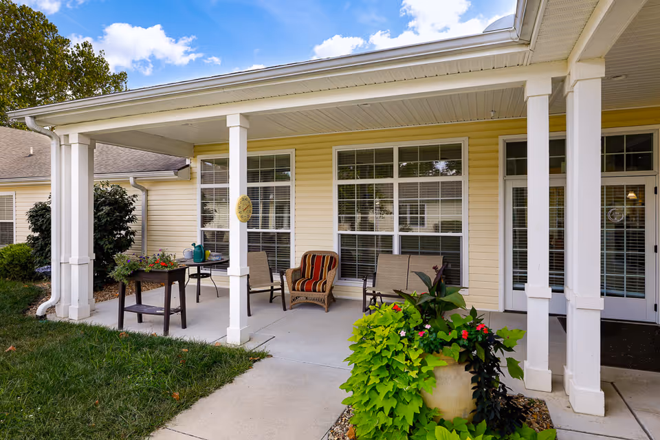 Covered outdoor patio area at Homestead Assisted Living of Emporia with several chairs, a small table with a watering can and potted plants, and a large planter with green and flowering plants near the entrance. The building exterior is light yellow with white trim and large windows.