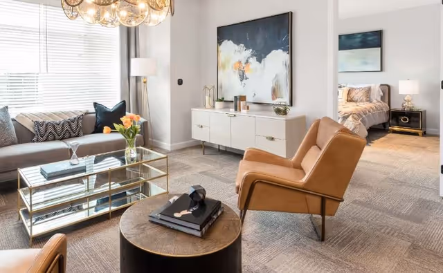 A modern living room with a beige sofa adorned with patterned pillows, a glass coffee table with books and a vase of orange flowers, two tan leather armchairs, a round wooden side table with books, a white sideboard with decorative items, and abstract artwork on the wall. In the background, a bedroom with a bed, nightstand, lamp, and artwork is visible through an open doorway.
