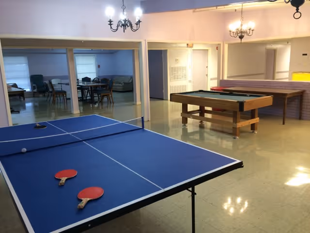 A recreational room featuring a blue ping pong table with two red paddles and a ball on it, a pool table, and a shuffleboard table. In the background, there is a seating area with chairs and tables, and the room is lit by chandeliers hanging from the ceiling.
