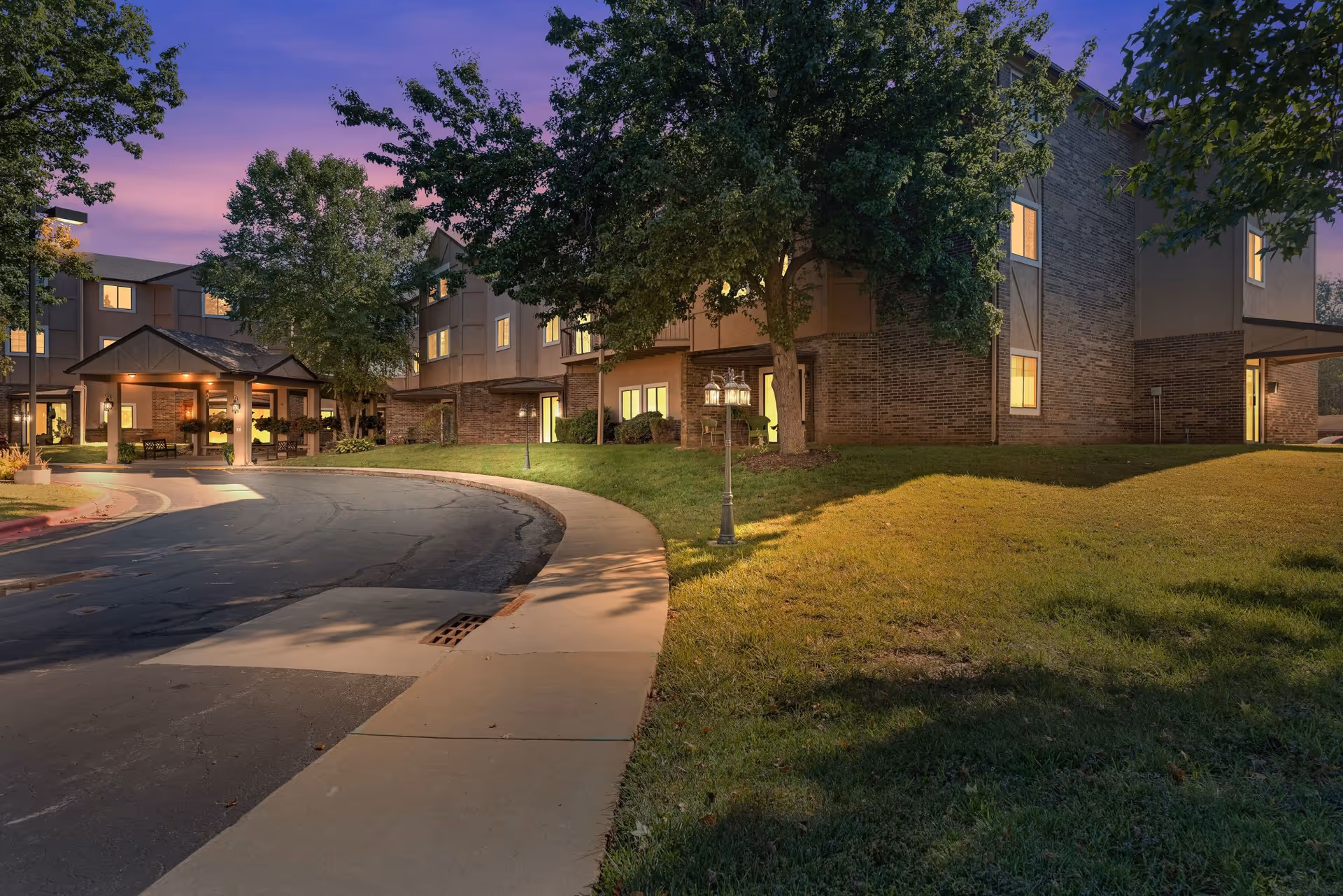 Dusk view of The Cambridge Senior Living building front with a curved driveway, entrance canopy, lit windows, and landscaped lawn.