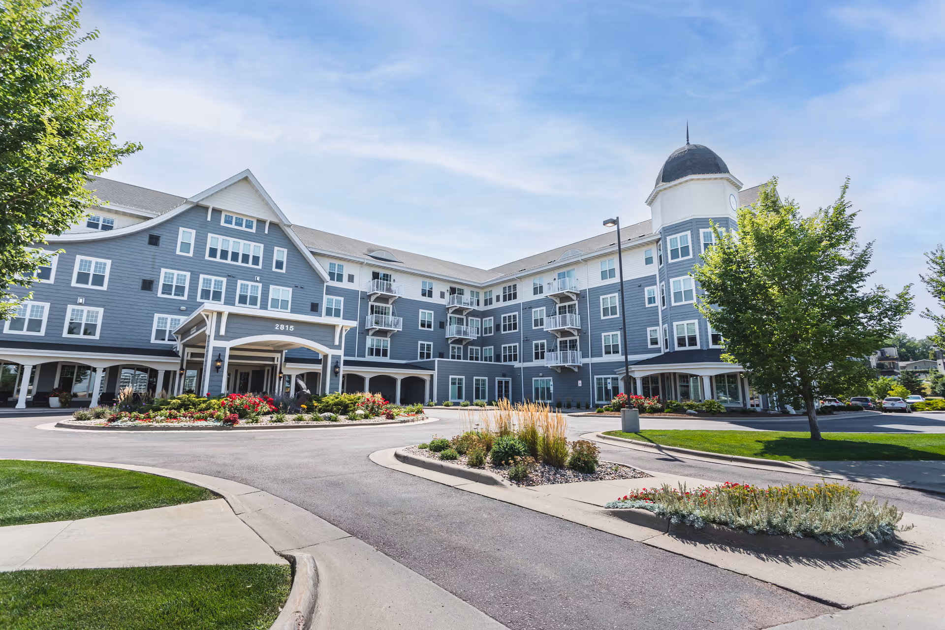 Exterior view of a large, multi-story senior living facility building with blue siding and white trim. The building features multiple windows, balconies, and a distinctive rounded tower with a dome roof. The entrance has a covered drop-off area with the number 2815 displayed. The surrounding area includes landscaped flower beds, trees, and a paved driveway under a clear blue sky.