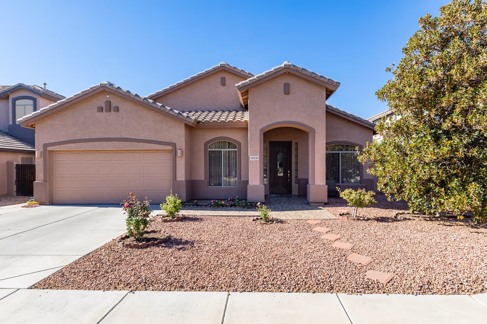Front exterior view of a single-story house with a tiled roof, two large windows with vertical blinds, a dark front door under an arched entryway, a two-car garage, and a landscaped front yard with gravel, small plants, and a large tree on the right side.