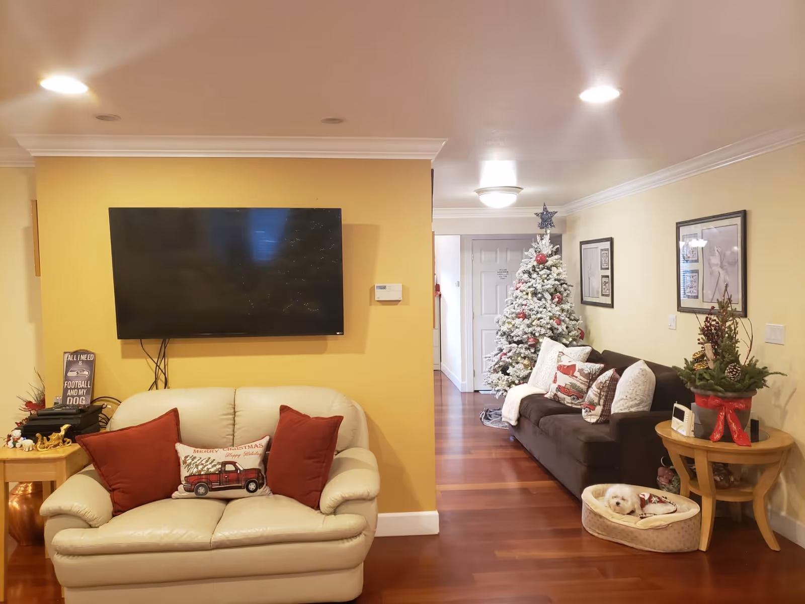 A cozy living room with a cream-colored loveseat adorned with red and Christmas-themed pillows, a wall-mounted flat-screen TV above it, and a dark brown sofa decorated with festive pillows next to a decorated Christmas tree. A small dog is resting in a pet bed on the wooden floor near a round wooden side table with holiday decorations.