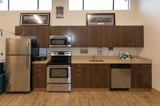 A modern kitchen area with stainless steel appliances including a refrigerator, microwave, stove, and dishwasher. The kitchen has wooden cabinets above and below the countertop, a sink, and some items on the counter such as a basket and a small decorative object. There are two framed pictures on the wall above the cabinets and a motivational sign between the pictures. The floor is wooden and the room is well-lit with natural light from windows above.