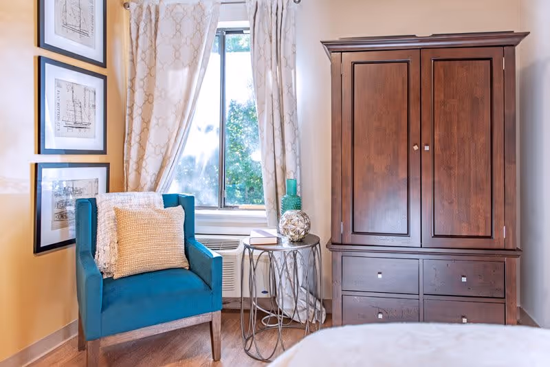 A cozy corner of a room featuring a teal upholstered armchair with a beige pillow and a white throw draped over the back. Next to the chair is a small round metal side table holding a decorative glass orb, a green vase, and a book. Behind the chair is a window with patterned white curtains, and to the right is a large wooden armoire with drawers at the bottom. Three framed architectural sketches hang on the wall to the left of the window.