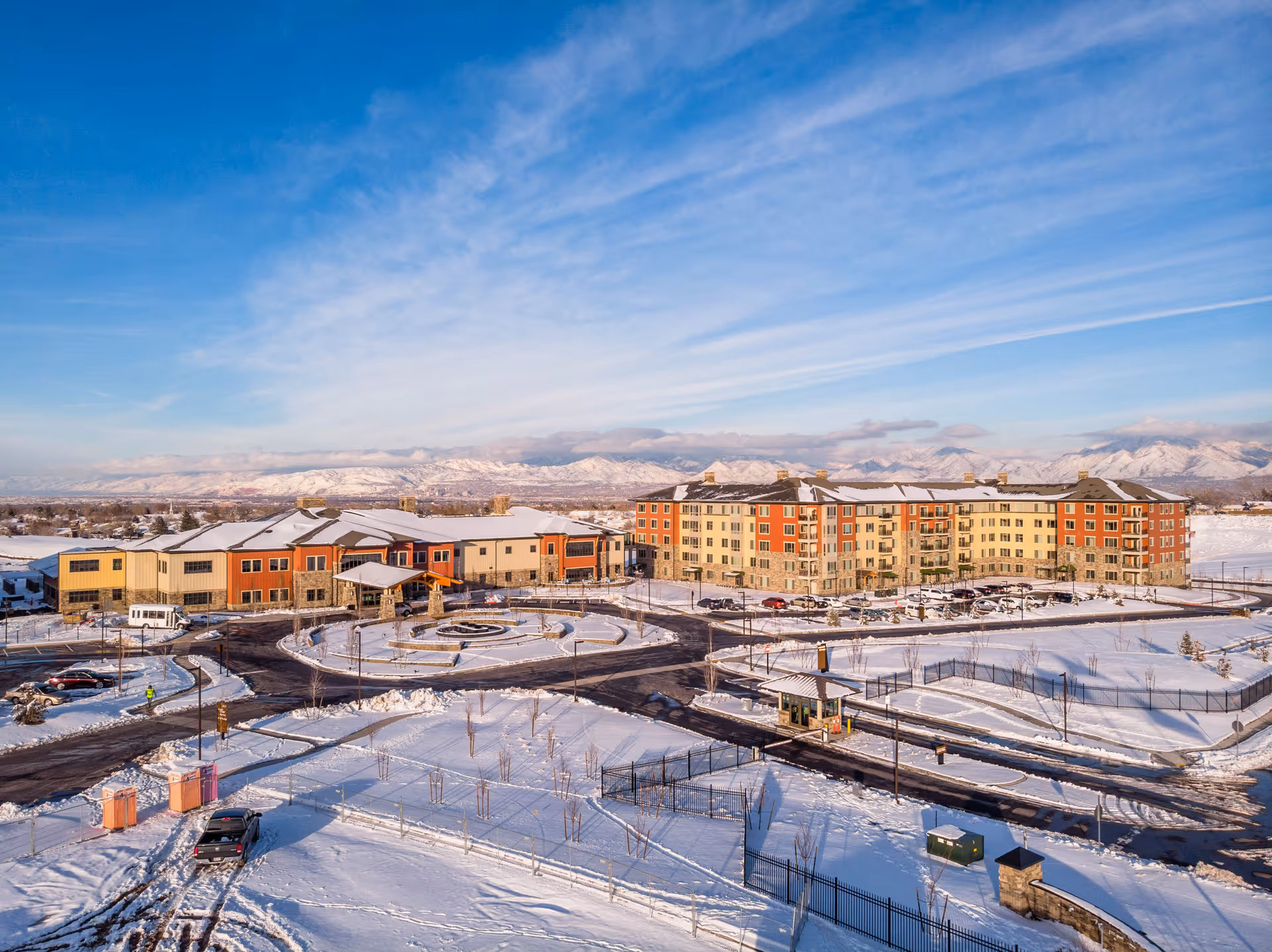 Aerial view of Summit Vista senior living facility in a snowy landscape with a large multi-story building and surrounding parking lots, roads, and fenced areas under a blue sky with scattered clouds and mountains in the background.