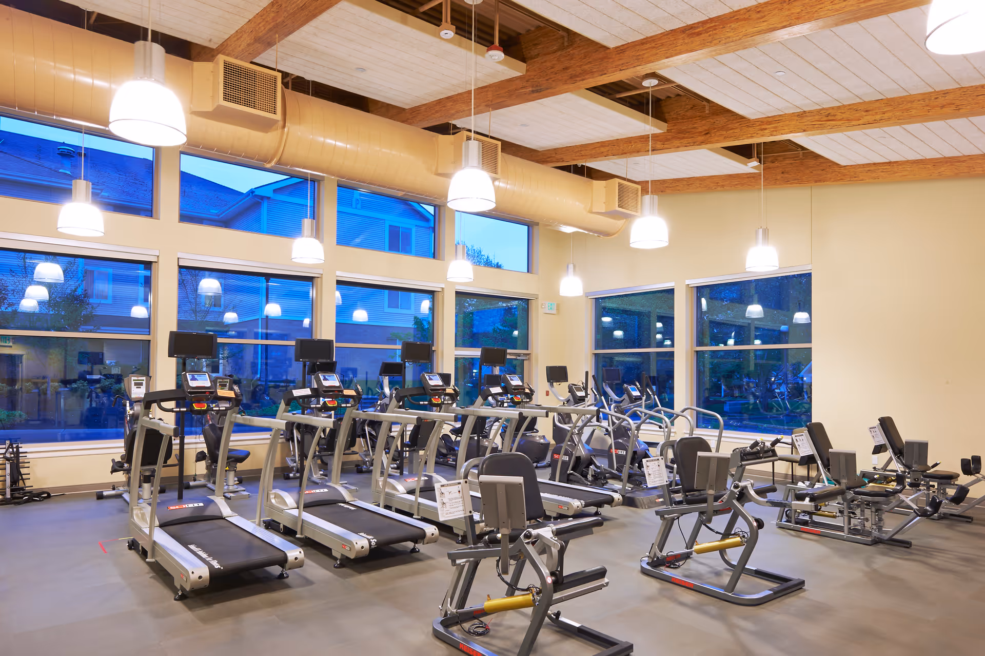 A spacious fitness room with multiple treadmills, exercise bikes, and other workout equipment. The room has large windows allowing natural light to enter, wooden ceiling beams, and several hanging pendant lights.