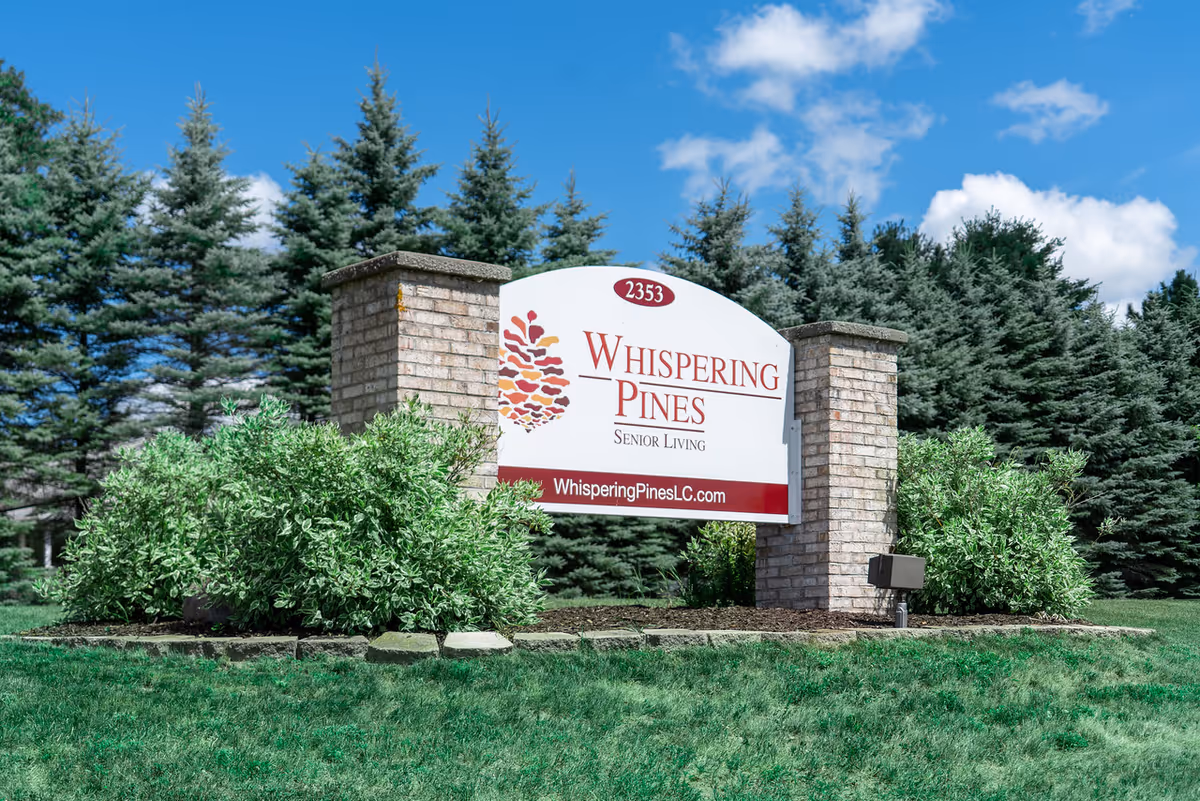 Entrance sign for Whispering Pines Senior Living between two brick pillars with shrubs and pine trees in the background under a blue sky.