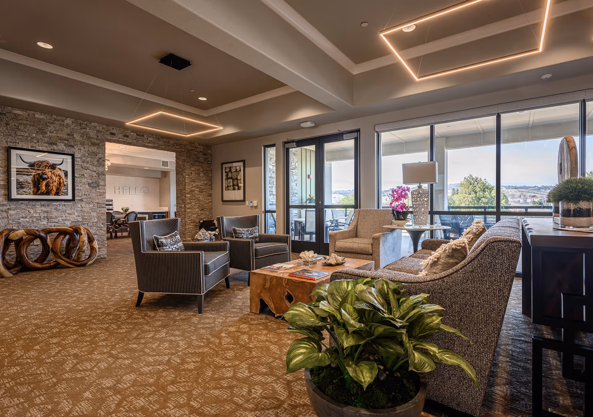 A spacious and well-lit living room area with modern furniture including two armchairs, a sofa, a wooden coffee table, and side tables with decorative items. Large windows and glass doors provide a view of an outdoor patio and landscape. The room features a stone accent wall with framed artwork, contemporary ceiling lights, and a patterned carpet. A large potted plant is in the foreground.