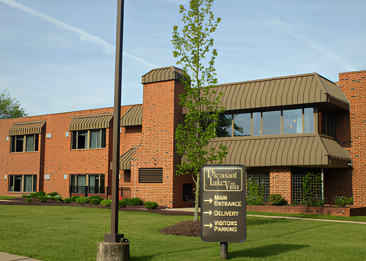 Brick two-story senior living building with brown metal awnings, a lawn, and a front sign reading 'Pleasant Lake Villa'.