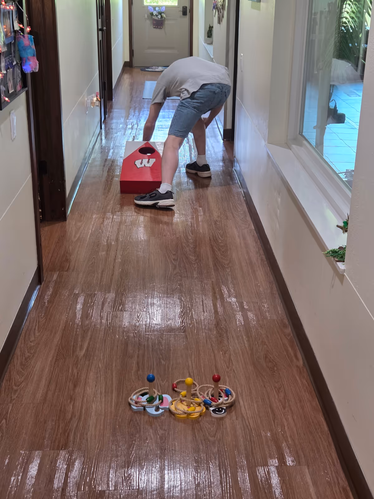 A person bending over in a hallway with wooden flooring, playing a game involving a red cornhole board with a white 'W' on it. There are three colorful ring toss targets placed on the floor in the foreground. The hallway has cream-colored walls, a window on the right side, and a door at the far end.