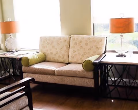 A cozy living room area with a beige patterned loveseat featuring green cylindrical pillows on each side. The loveseat is flanked by two dark wood side tables, each with an orange lampshade and decorative items. Large windows behind the loveseat allow natural light to brighten the room.