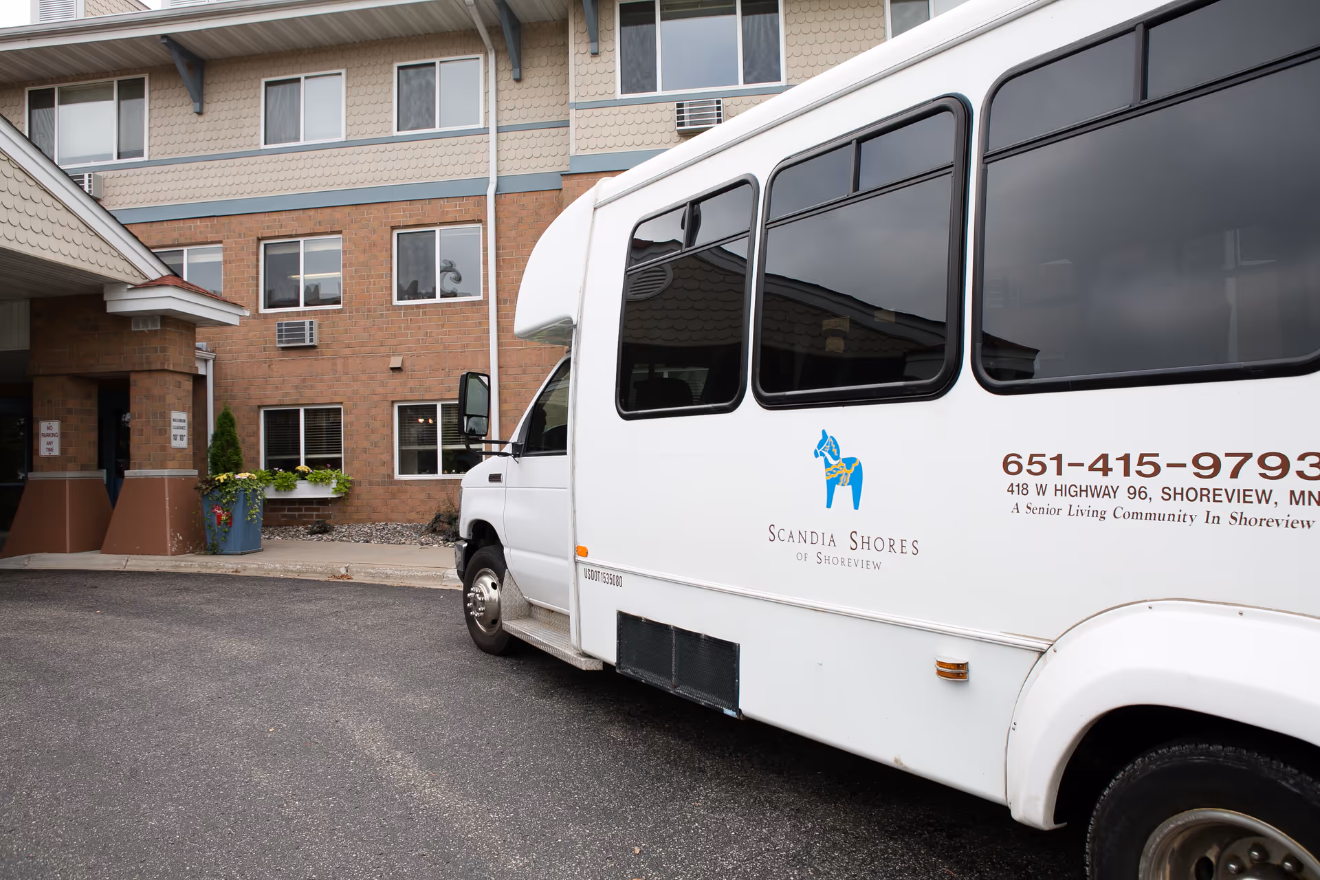 A white shuttle bus parked outside a brick and beige senior living facility building. The bus has the logo and name 'Scandia Shores of Shoreview' along with contact information and address on its side. The building has multiple windows and a covered entrance with some plants near the doorway.