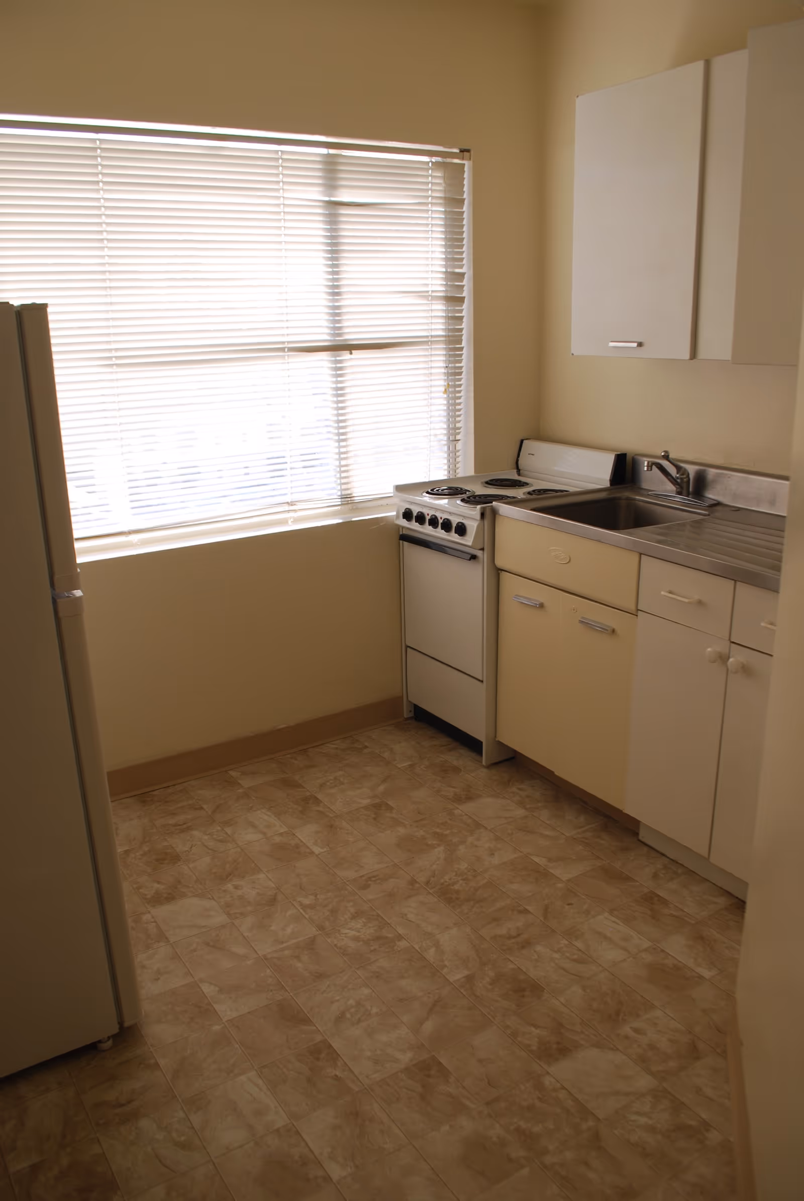 Small kitchen with a window and blinds, a compact electric stove, sink, and light-colored cabinets.