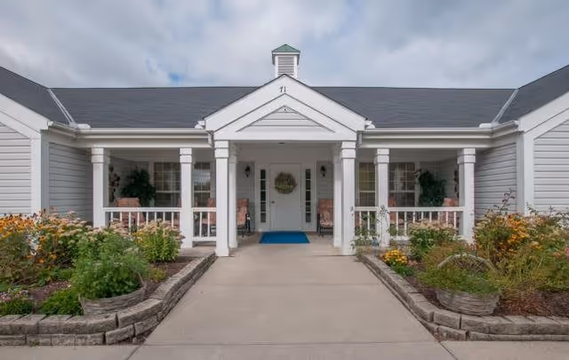 Front entrance of a single-story building with white columns and a covered porch. There are flower beds with various plants and flowers on either side of the concrete walkway leading to the entrance. The building has gray siding and a dark shingled roof under a cloudy sky.
