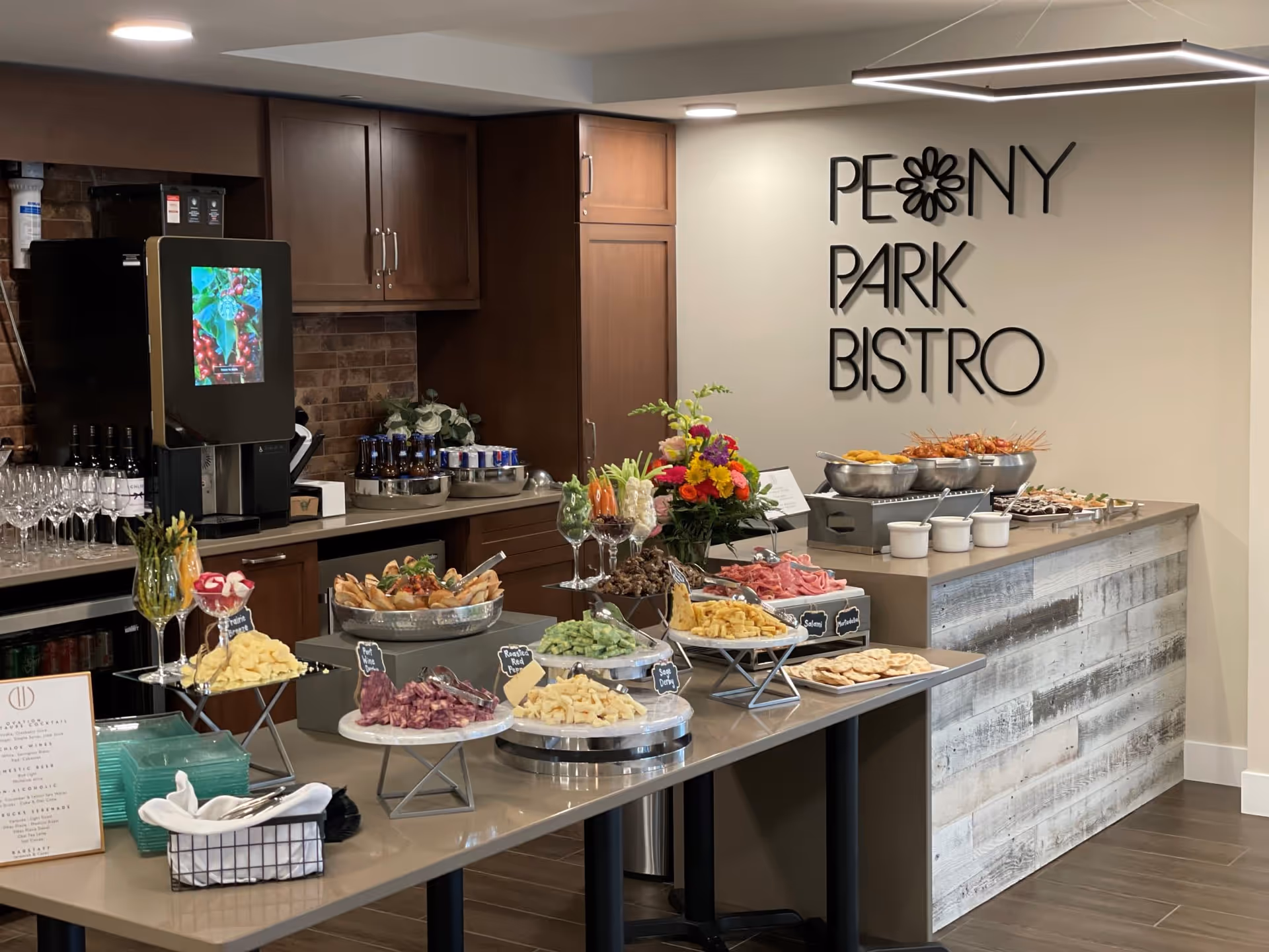 Interior view of a dining area named Peony Park Bistro featuring a buffet setup with various appetizers including cheeses, meats, vegetables, and crackers. The space has wooden cabinetry, a beverage dispenser, and a rustic wooden counter with bowls of food. A colorful flower arrangement is placed on the counter.