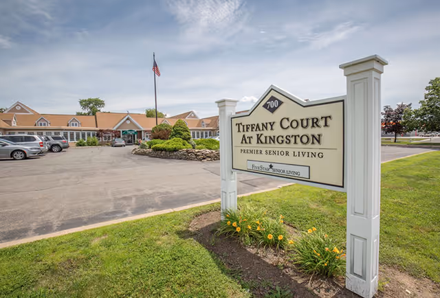 Sign reading 'Tiffany Court at Kingston' in front of the senior living facility building, parking lot, and landscaped entrance.